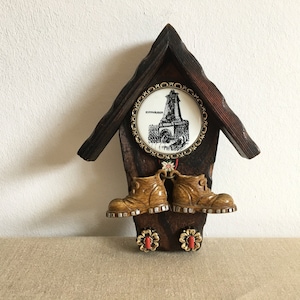 May include: A wooden wall hanging shaped like a house with a round, white plaque that says "Kyffhäuser" and a black and white image of a castle. Two brown, plastic boots hang from the bottom of the house.