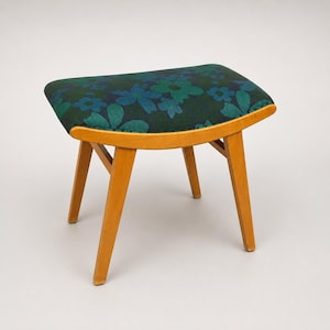 Può includere: Uno sgabello piccolo con quattro gambe in legno chiaro e una struttura in legno curva. La seduta è rivestita in un tessuto scuro con un motivo ripetuto di grandi fiori stilizzati in varie tonalità di blu e verde su sfondo scuro.