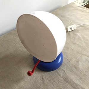 May include: A white table lamp with a blue base and a red handle. The lamp has a round, white shade with a pattern of small holes.
