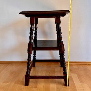 May include: A dark brown wooden side table with a square top and a lower shelf. The table features four turned legs with decorative block accents. A yellow measuring tape is positioned vertically next to the table, indicating a height of 31 inches.