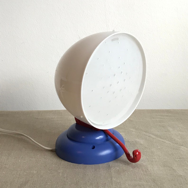 May include: A white lamp with a blue base and a red cord. The lamp has a round, perforated shade and a small handle.