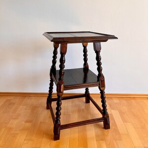 May include: A dark brown wooden two-tier side table featuring a square top and a lower square shelf. The table is supported by four turned legs with decorative block accents at the base and mid-section, showcasing a polished wood grain finish.