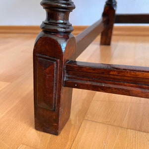 May include: A dark brown wooden furniture leg with a turned, rounded top section and a square base featuring a recessed rectangular panel. A horizontal wooden support beam connects to the leg, showcasing a polished finish and visible wood grain.
