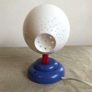 May include: A white sphere lamp with a blue and red base. The sphere has a pattern of small holes and a larger hole in the center. The lamp has a white cord.