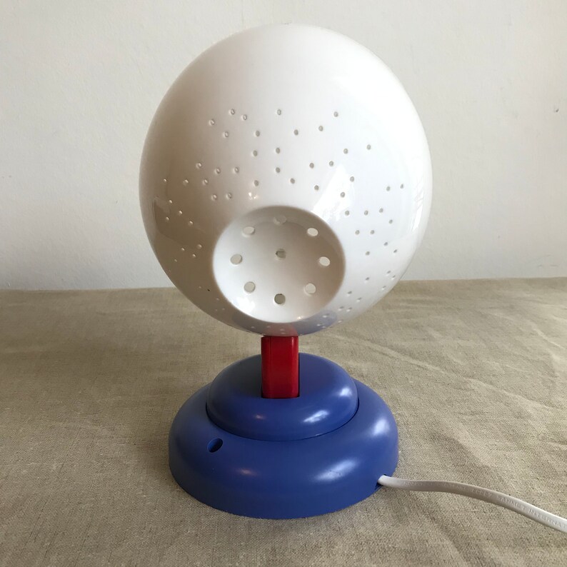 May include: A white sphere lamp with a blue and red base. The sphere has a pattern of small holes and a larger hole in the center. The lamp has a white cord.