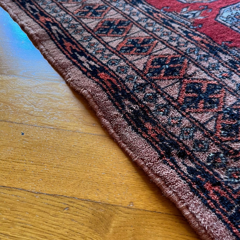 May include: Close-up of a rug with a detailed geometric pattern in shades of red, black, and beige. The rug has a fringed edge.