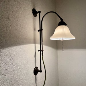 Peut inclure: Une lampe murale réglable de couleur bronze avec un abat-jour en verre dépoli blanc. La lampe est équipée d'un interrupteur à tirette et est montée sur un mur blanc.