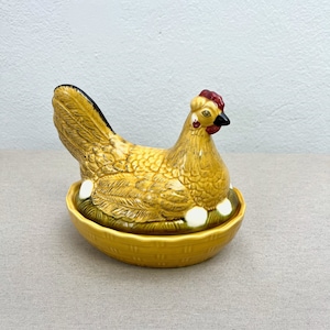 Könnte beinhalten: Eine Keramik-Hühnerfigur mit gelbem Körper, braunen Flügeln und einem roten Kamm. Das Huhn sitzt auf einem Nest mit weißen Eiern.