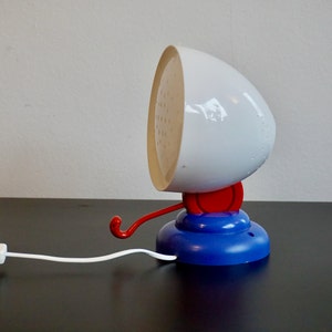 May include: A white and blue table lamp with a red switch and a hook. The lamp has a white, round shade with a textured surface.
