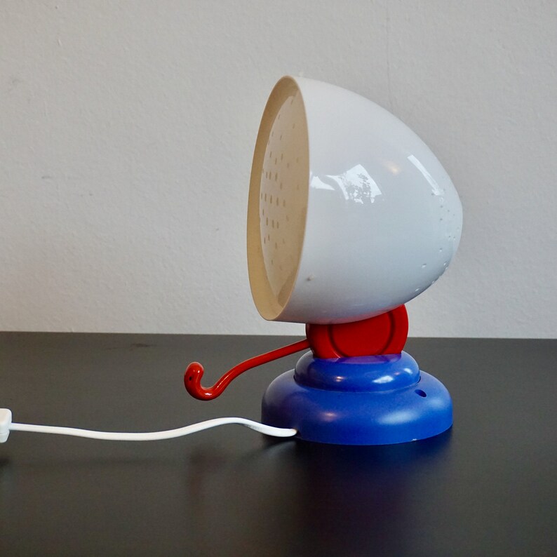 May include: A white and blue table lamp with a red switch and a hook. The lamp has a white, round shade with a textured surface.