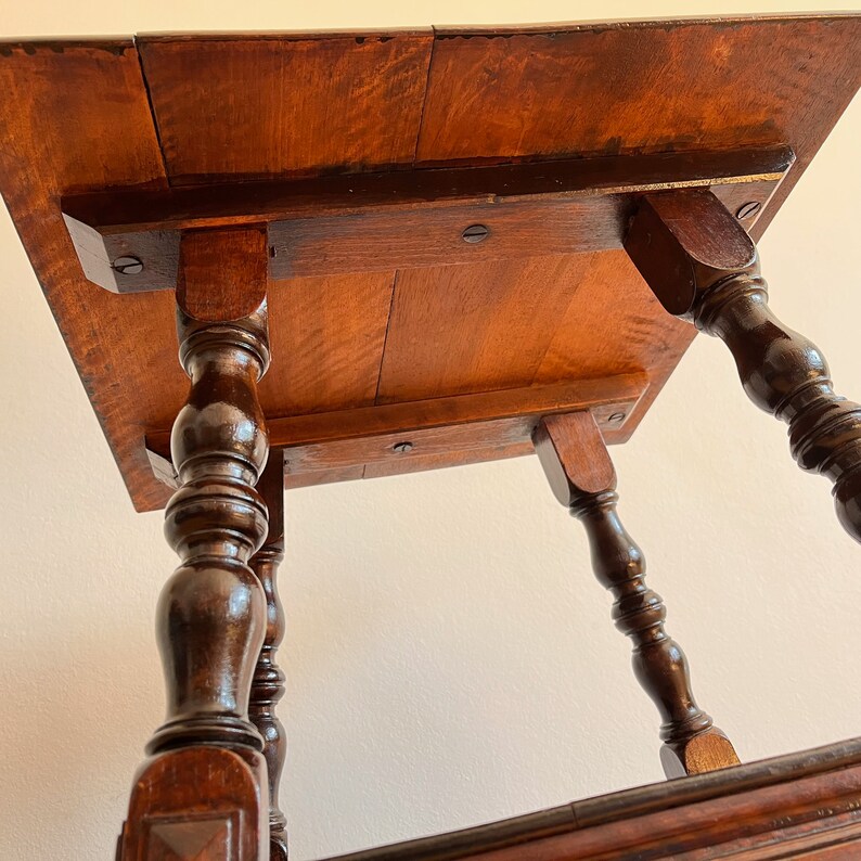 May include: A dark brown wooden side table featuring a square top with visible wood grain and four turned, spindle-style legs. The underside of the tabletop is reinforced with horizontal wooden supports secured by dark metal screws.