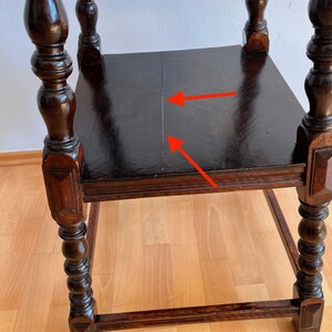 May include: A dark brown wooden side table with turned legs and a square top surface. The top surface features a visible vertical seam running down the center, highlighted by two red arrows pointing toward the gap in the wood grain.