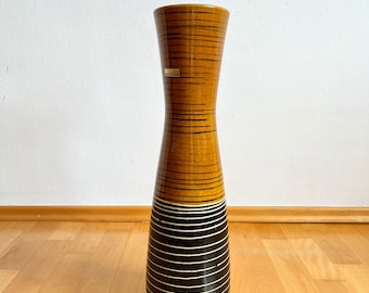 May include: A tall, hourglass-shaped ceramic floor vase featuring a glossy finish. The upper section is a warm ochre yellow with thin horizontal black lines, while the lower section is dark brown with thin horizontal white stripes.