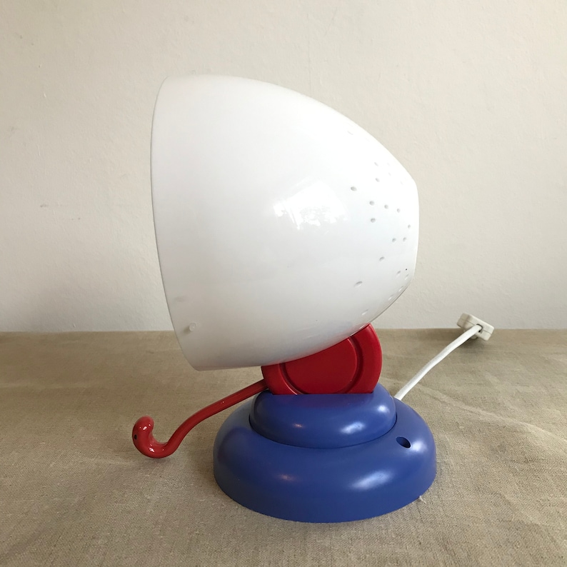 May include: A white, round lamp with a red and blue base. The lamp has a cord and a hook on the side.