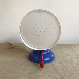 May include: A white lamp with a blue base and a red handle. The lamp has a white shade with a pattern of small holes.