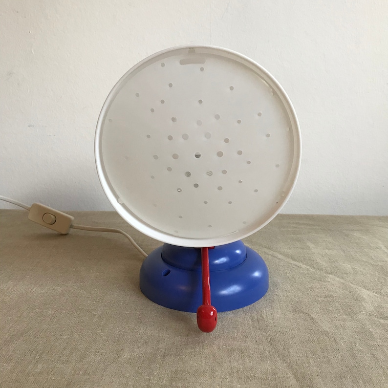May include: A white lamp with a blue base and a red handle. The lamp has a white shade with a pattern of small holes.