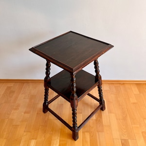 May include: A dark brown wooden side table featuring a square top and a lower square shelf. The table is supported by four turned legs with decorative carved details and block feet, showcasing a polished finish and visible wood grain throughout.