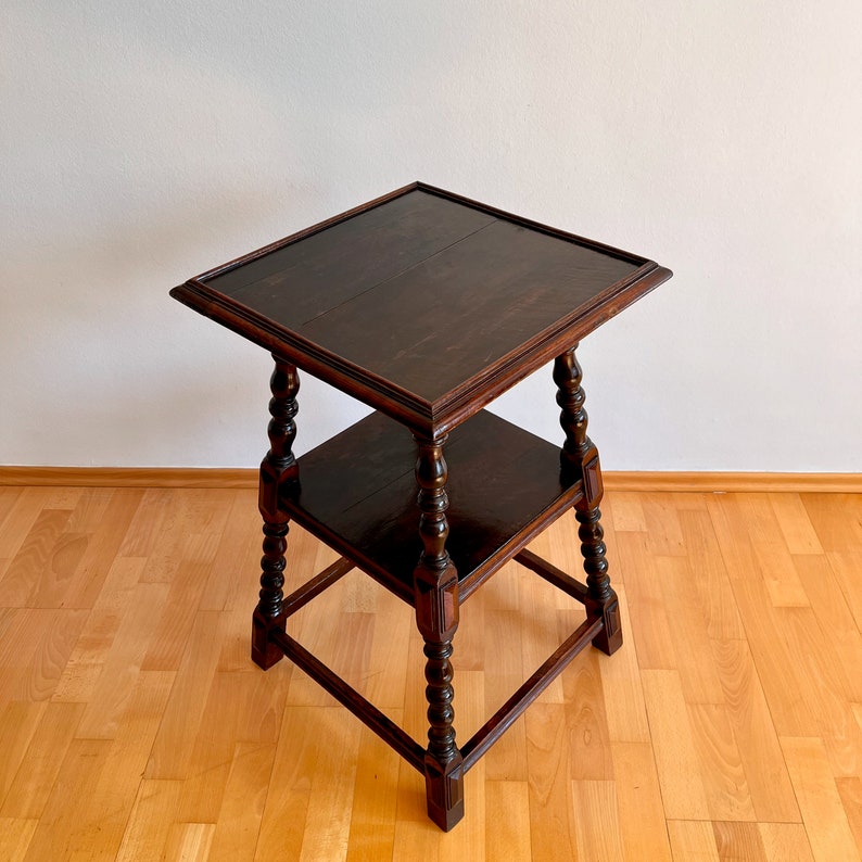 May include: A dark brown wooden side table featuring a square top and a lower square shelf. The table is supported by four turned legs with decorative carved details and block feet, showcasing a polished finish and visible wood grain throughout.