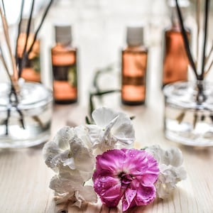 May include: Three glass bottles with brown liquid and black sticks in them, sitting on a wooden surface.  A bouquet of white and pink flowers sits in front of the bottles.