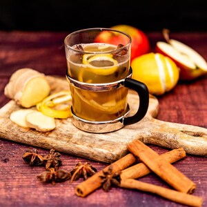 Pode incluir: Uma caneca de vidro cheia de uma bebida quente, possivelmente sidra temperada ou sidra de maçã, com fatias de limão e paus de canela. A caneca está sobre uma tábua de cortar de madeira com fatias de gengibre, anis estrelado e paus de canela.