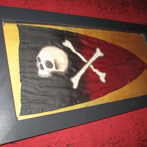 Può includere: Una bandiera pirata nera e rossa incorniciata con un design di teschio e ossa incrociate bianche. La bandiera è esposta in una cornice nera con un passepartout dorato.