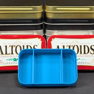 May include: A collection of Altoids mint tins in gold, black, and silver, with a blue organizer in front. The Altoids tins are red and white with text. The blue organizer has three compartments.