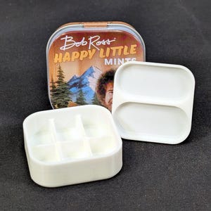 Puede incluir: Una lata de metal con el texto "Bob Ross HAPPY LITTLE MINTS" y una imagen de Bob Ross y una escena de montaña. La lata está abierta, revelando insertos de plástico blanco con dos y seis compartimentos.
