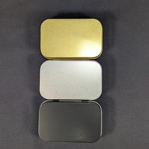May include: Three small, rectangular metal tins in gold, silver, and black. The tins are arranged vertically, with the gold tin at the top, the silver in the middle, and the black at the bottom. The tins have rounded corners and a hinged lid.
