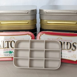 May include: Two Altoids mint tins, one red and one silver, with the text "Original Celebrated Altoids Curiously Strong Peppermints" and "The Original Celebrated Altoids Strong Mints" on the front. A white plastic tray with nine compartments is in front of the tins.