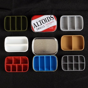 May include: A collection of eight small, rectangular containers with various colours and compartments. The containers are made of silicone and are designed to hold small items, such as pills, jewellery, or craft supplies. The containers are arranged in two rows of four, with the top row featuring a green container, a silver tin with the text "Altoids Smalls Peppermint" on the lid, a grey container, and a brown container. The bottom row features a white container, a blue container, a grey container, and a red container.