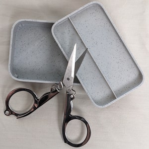 May include: A silver folding scissors with a gray speckled plastic case. The case has a divider in the middle.