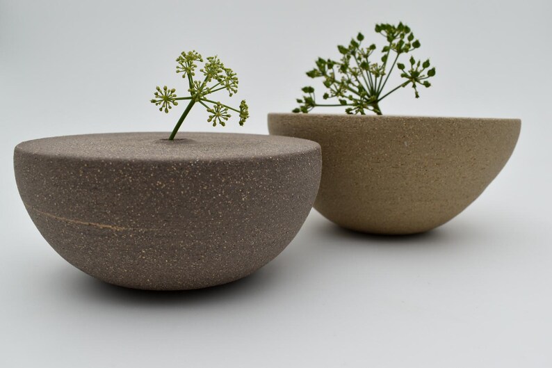 May include: Two contemporary ceramic bowls in brown and beige, each with a sprig of green foliage. The bowls have a minimalist design, ideal for displaying small plants or flowers. The bowls are measured in centimetres.