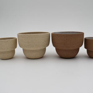 Op de afbeelding: Vier keramische potten in verschillende maten en kleuren. Twee zijn lichtbeige met een textuuroppervlak en twee zijn donkerbruin. Elke pot heeft een afgeronde basis en een bredere, ovaalvormige bovenkant.