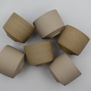 Puede incluir: Seis tazas de cerámica hechas a mano en tonos beige y marrón se disponen sobre una superficie blanca. Las tazas presentan un diseño angular único con un acabado texturizado, mostrando una estética minimalista.