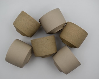 May include: Six handmade ceramic cups in shades of beige and tan are arranged on a white surface. The cups have a unique, angled design with a textured finish, showcasing a minimalist aesthetic. These cups are ideal for coffee or tea.