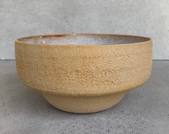 May include: A large, round, ceramic bowl with a textured, sandy brown finish. The bowl has a slightly flared rim and a rounded base.