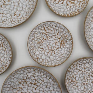 Puede incluir: Colección de siete cuencos de cerámica blanca con un patrón texturizado, similar a un mosaico. Los cuencos tienen un borde marrón claro y están dispuestos sobre una superficie blanca. Los cuencos son redondos y de diferentes tamaños.