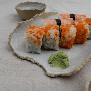 Può includere: Un piatto in ceramica bianca con una superficie ruvida e testurizzata contiene diversi rotoli di sushi. I rotoli sono ricoperti di uova di pesce arancioni e c'è una pasta di wasabi verde sul piatto.