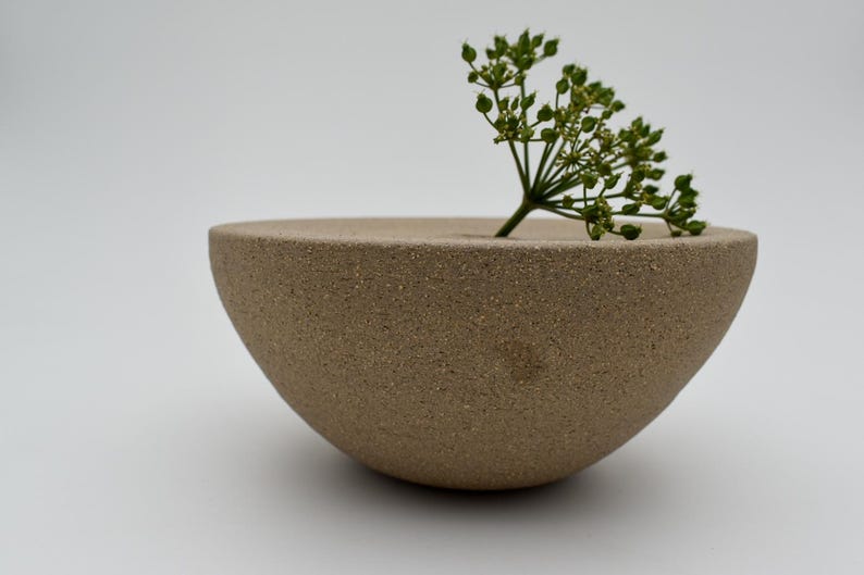 May include: A beige, semi-spherical ceramic vase with a small opening, holding a sprig of green plant. The vase has a textured surface and sits on a white background. A minimalist, modern design.