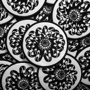 May include: A close-up of several black and white embroidered patches featuring a stylized flower design. The patches are circular and have a white background with a black outline.