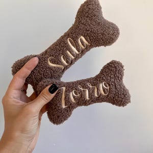 Può includere: Due giocattoli per cani a forma di osso marrone con nomi ricamati. Uno dice "Sula" e l'altro "Zorro" in corsivo. I giocattoli hanno un aspetto morbido e strutturato, su sfondo bianco.