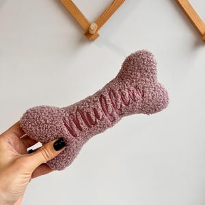 May include: A soft, mauve dog bone toy with the name "Mullu" embroidered in a darker pink thread. The toy has a textured, fuzzy surface and is shaped like a bone. A pet accessory.