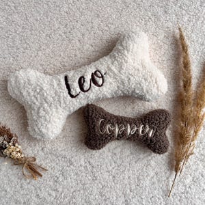 Könnte beinhalten: Zwei Plüsch-Hundespielzeuge in Form eines Knochens und einer Schleife. Das knochenförmige Spielzeug ist weiß mit dem Namen "Leo" in Dunkelbraun gestickt. Das Schleifenspielzeug ist braun mit "Copper" in Gold gestickt.