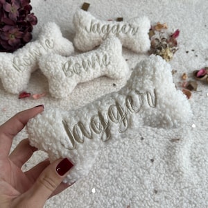 Può includere: Giocattoli per cani a forma di osso bianco con nomi ricamati. Uno è tenuto in mano. I nomi "Jagger" e "Bowie" sono ricamati sui giocattoli. I giocattoli sono su una superficie bianca strutturata con accenti floreali.