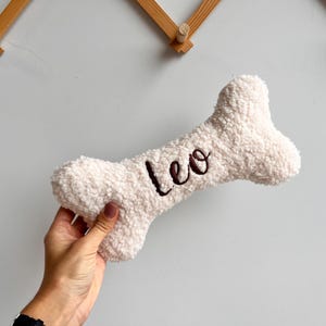 Könnte beinhalten: Ein cremefarbenes, knochenförmiges Hundespielzeug. Das Wort "Leo" ist in dunkelbrauner Schrift über die Mitte gestickt. Das Spielzeug hat ein weiches, strukturiertes Aussehen, was auf ein komfortables und langlebiges Design für Haustiere hindeutet.