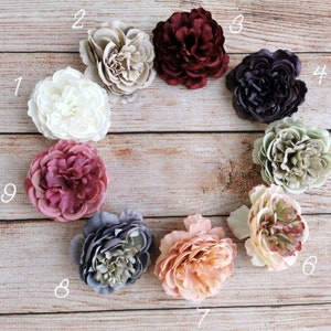 Puede incluir: Nueve cabezas de flores artificiales en varios colores, incluyendo blanco, beige, burdeos, morado oscuro, verde salvia, melocotón y gris. Las flores están dispuestas en círculo sobre una superficie de madera.