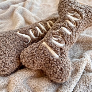 May include: Two brown, bone-shaped dog toys with the word "Love" embroidered in white. The toys are made of a soft, textured material and rest on a plush, tan blanket. A cozy pet accessory.