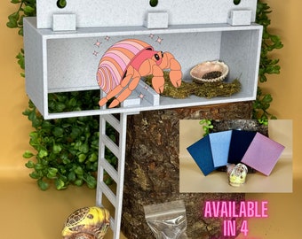 May include: A light grey hermit crab habitat with a ladder, a cartoon hermit crab, and a seashell. The text "AVAILABLE IN 4 COLORS!" is displayed. Includes a bag of suction cups and four fabric swatches.