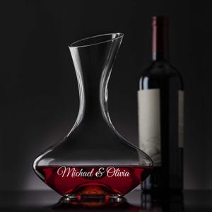 Puede incluir: Un elegante decantador de vino de vidrio transparente lleno de vino tinto, con los nombres "Michael & Olivia" elegantemente escritos en la parte delantera. Una botella de vino oscuro con una etiqueta en blanco se encuentra en el fondo, sobre un fondo oscuro.