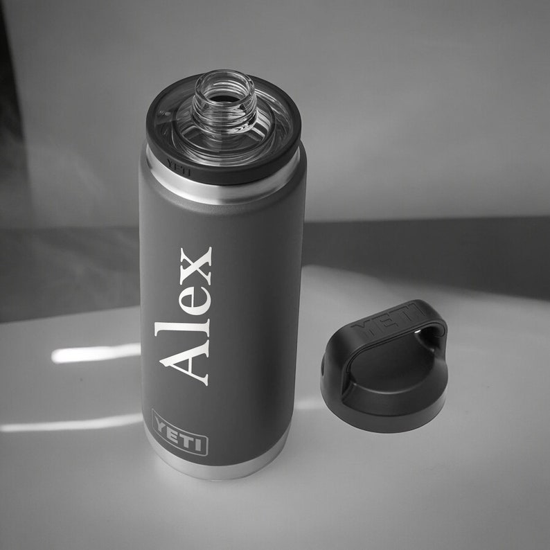 Puede incluir: Una botella de agua aislada de color gris oscuro con el nombre "Alex" en letras blancas. La botella tiene una tapa negra y el logotipo de YETI. La tapa est&aacute; a un lado.